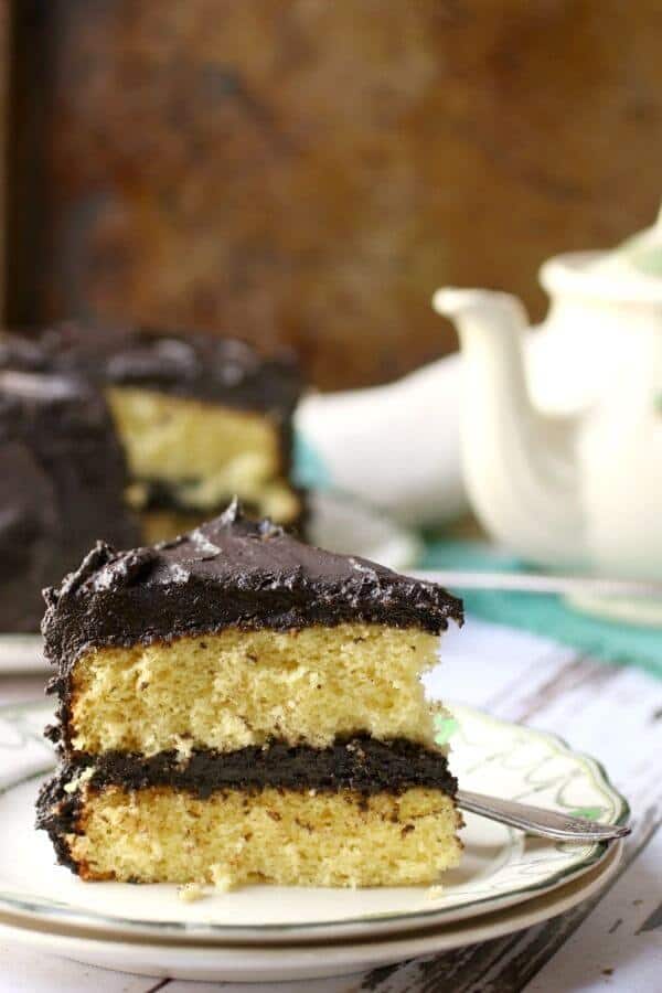 Yellow butter cake with fudge frosting on a painted table with a teapot in the background