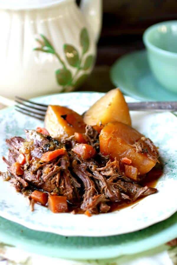 Perfect pot roast with au jus, potatoes, and carrots are served up on a vintage white and green plate. A tea pot is in the background.