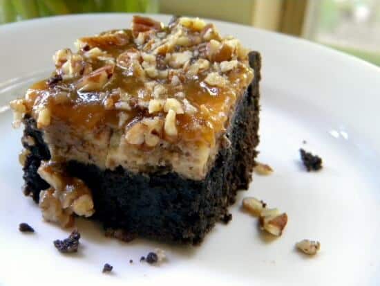 A brownie cheesecake bar on a plate with pecans tumbling off of it