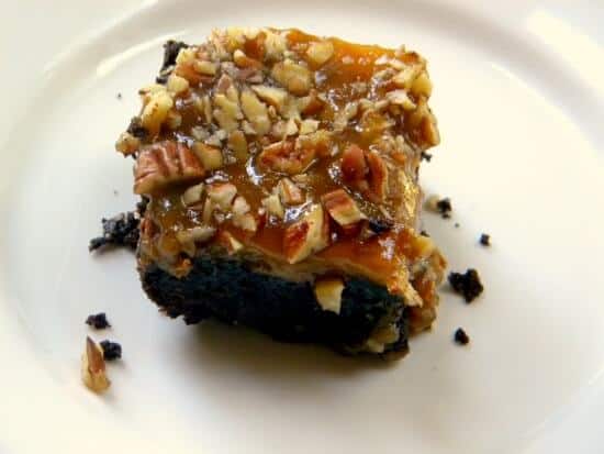 praline-cheesecake-brownies brownie cheesecake bars - a layered bar made of brownie,cheesecake, and pecan praline on a white plate