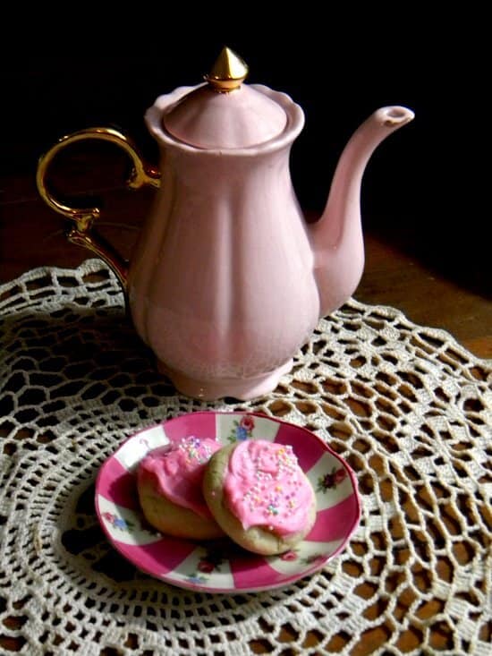 A pink teapot on a lace tablecloth with pink frosted cookies on a plate.