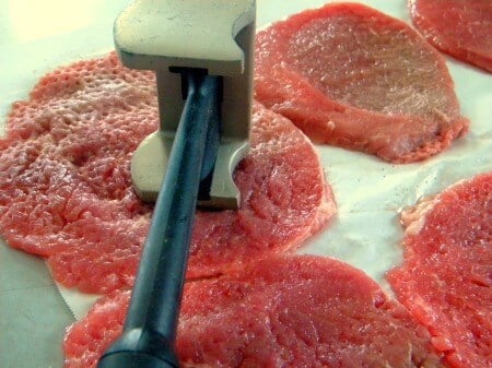 A raw round steak being tenderized with a mallet.