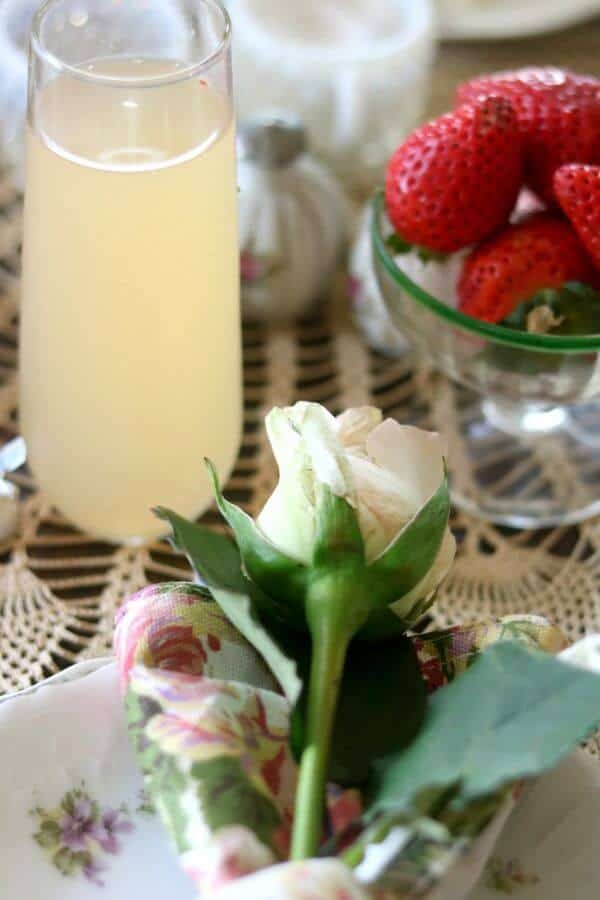 A grapefruit mimosa and dish of strawberries on a lace table cloth. A white rose is in the foreground.