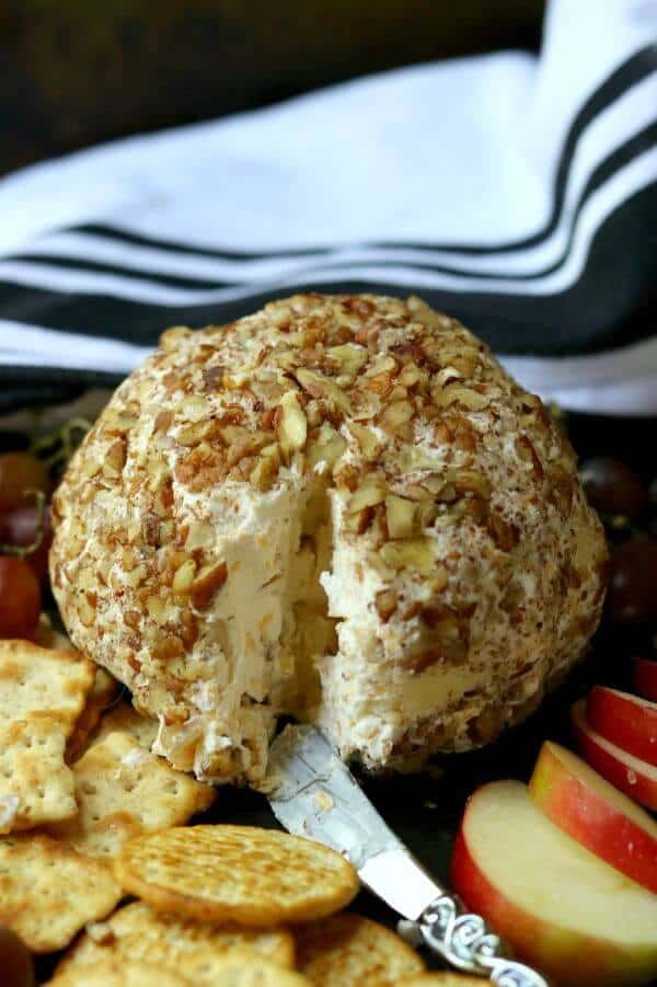 Apple cheddar cheeseball covered in pecans with a wedge cut out of it. Crackers and sliced apples are near by.