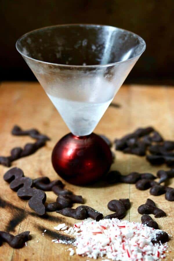 An empty glass, frosted from the freezer, waits to be filled with chocolate peppermint martinie. The glass is surrounded by chocolate squiggles and crushed peppermint.