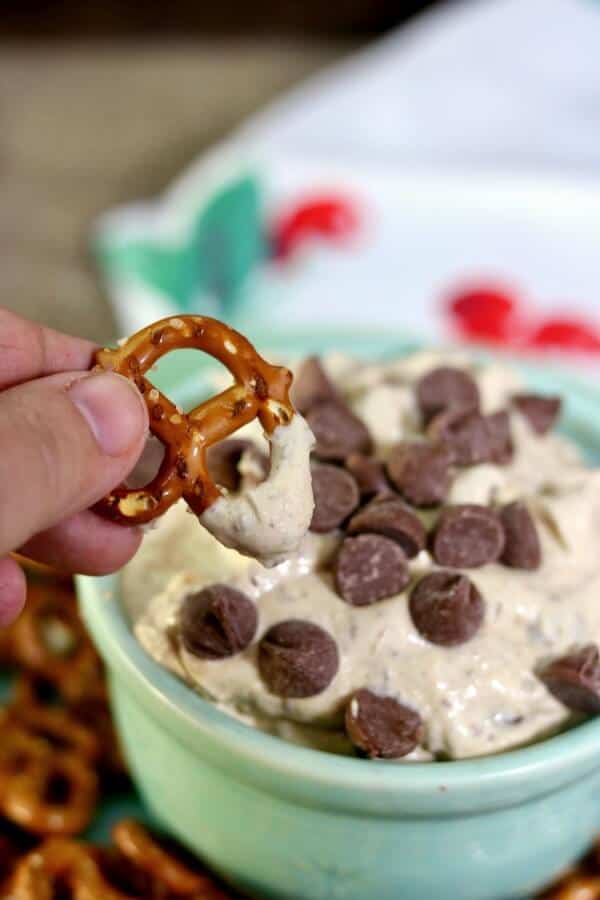 A hand dipping a pretzel into peanut butter chocolate chip cheesecake dip, which is in a green bow,.