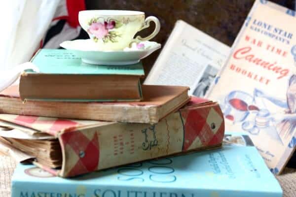 Feature image for best gifts for cookbook collectors. Stack of antique cookbooks with a vintage teacup and saucer on the top. Teacup has pink roses on it.