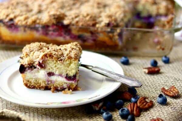 Blueberry cream cheese coffee cake square on a white plate with a silver fork. You can clearly see the layers of blueberries, cheese, and cake.