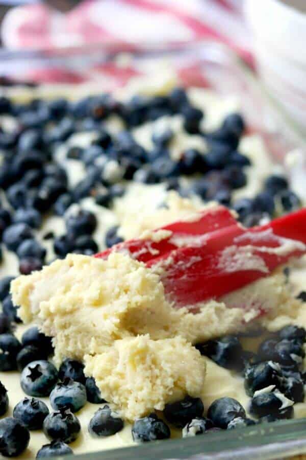 Topping being put on the blueberry cream cheese coffee cake with a red rubber spatual.