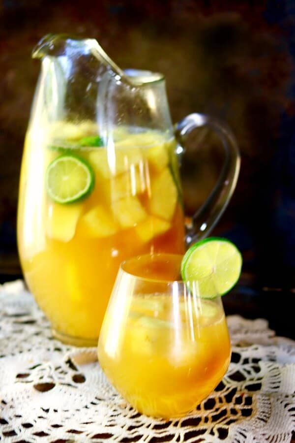 A glass pitcher of tropical pineapple sangria is behind a glass of sangria garnished with a lime.