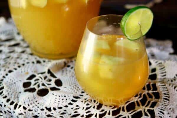 A glass of bright tropical pineapple sangria, garnished with a lime slice sits on a white lace cloth. Behind the glass is a pitcher of the sangria.