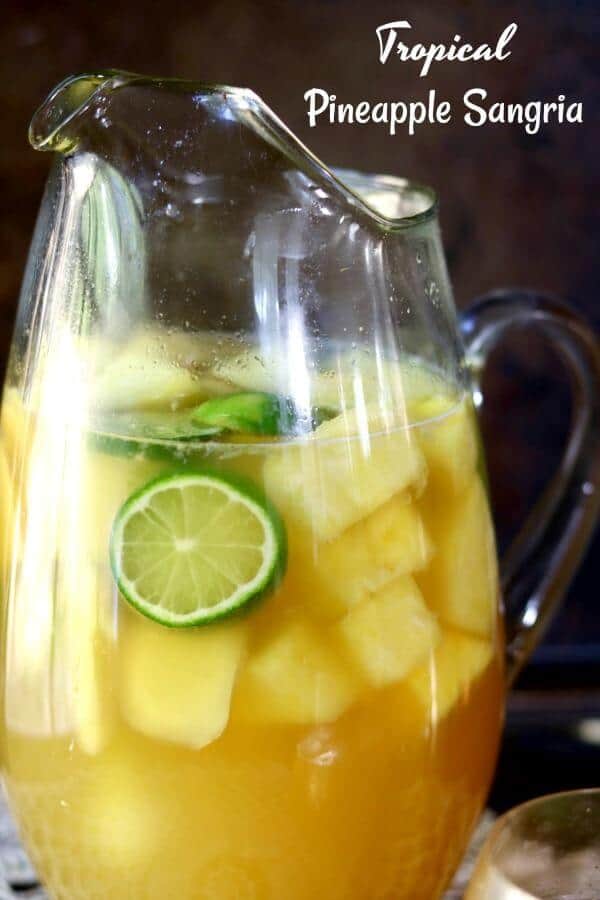 Closeup of a glass pitcher filled with yellow tropical pineapple sangria. Sliced of lime and bits of pineapple float in the pitcher.