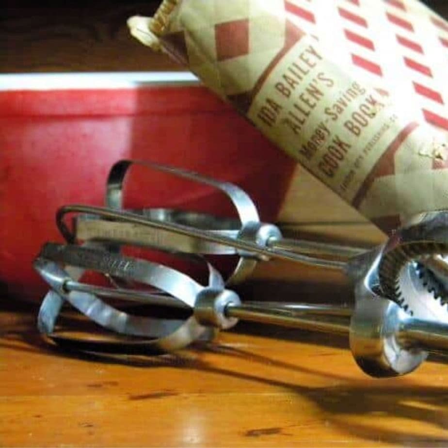 antique egg beater, cookbook and red mixing bowl on a wooden counter.