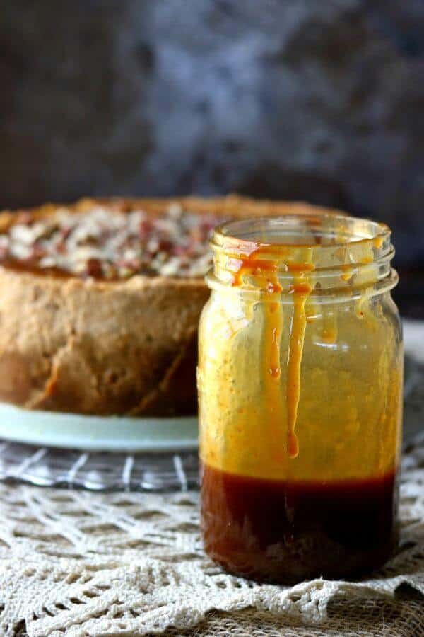 A jar of homemade caramel syrup is placed in front of a whole pumpkin cheesecake with gingersnap crust.