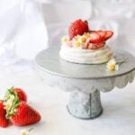 A small strawberry Pavlova is placed on a cake plate. Three strawberries are in the foreground.