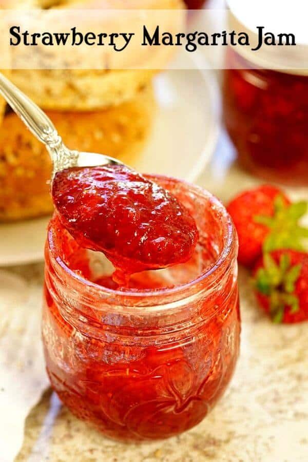A spoonful of strawberry jam is lifted out of a jar - ripe strawberries are in the background.