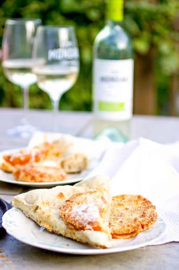 Using a pizza stone on the grill makes this a quick easy dinner- make this fried green tomato pizza with smoky cheese and whole wheat crust! #restlesschipotle #mondavi #recipe #howtomake #howtocook #homemade Slices of fried green tomatoes with glasses of white wine in the background.
