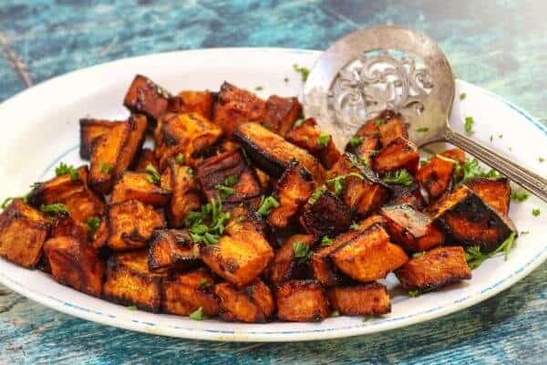 An antique platter of golden roasted sweet potatoes with a silver serving spoon