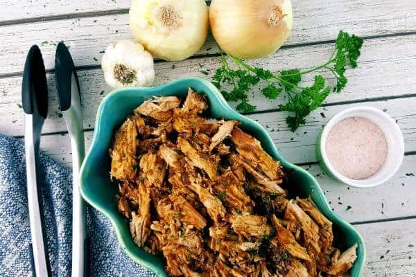 Pulled pork in an oval serving dish surrounded by garlic, onions, fresh herbs and salt.