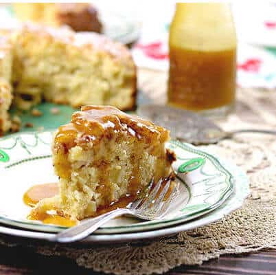 A thick slice of Irish Apple Cake is covered in brown butter whiskey sauce. The whole cake and the sauce in a bottle are in the background.