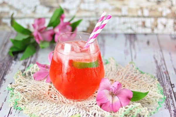 A red cocktail is on a piece of lace and surrounded by pink flowers.