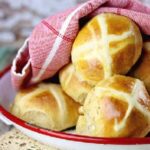 Golden, baked hot cross buns on a tray with a red tea towel over them.