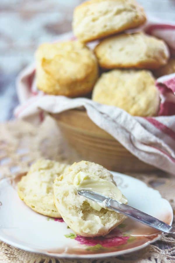 Butter being spread on an open biscuit - bowl of biscuits in the background.