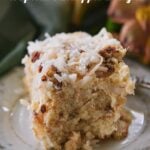 A square of pineapple coconut layered cake on a white plate with a green , pink, and gold floral edge. Orange blossoms in the background.