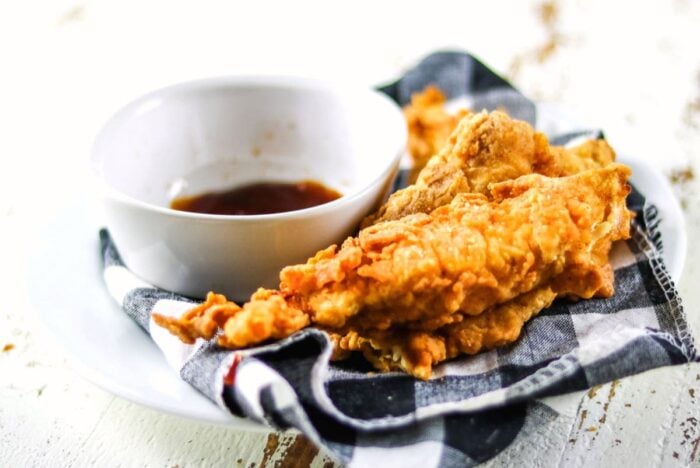 Finished Popeye's copycat chicken strips recipe on a plate next to a dish of bbq sauce.