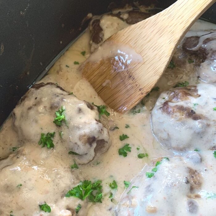 Finished hamburger steaks in the slow cooker covered with gravy. A wooden spoon is resting on the side.