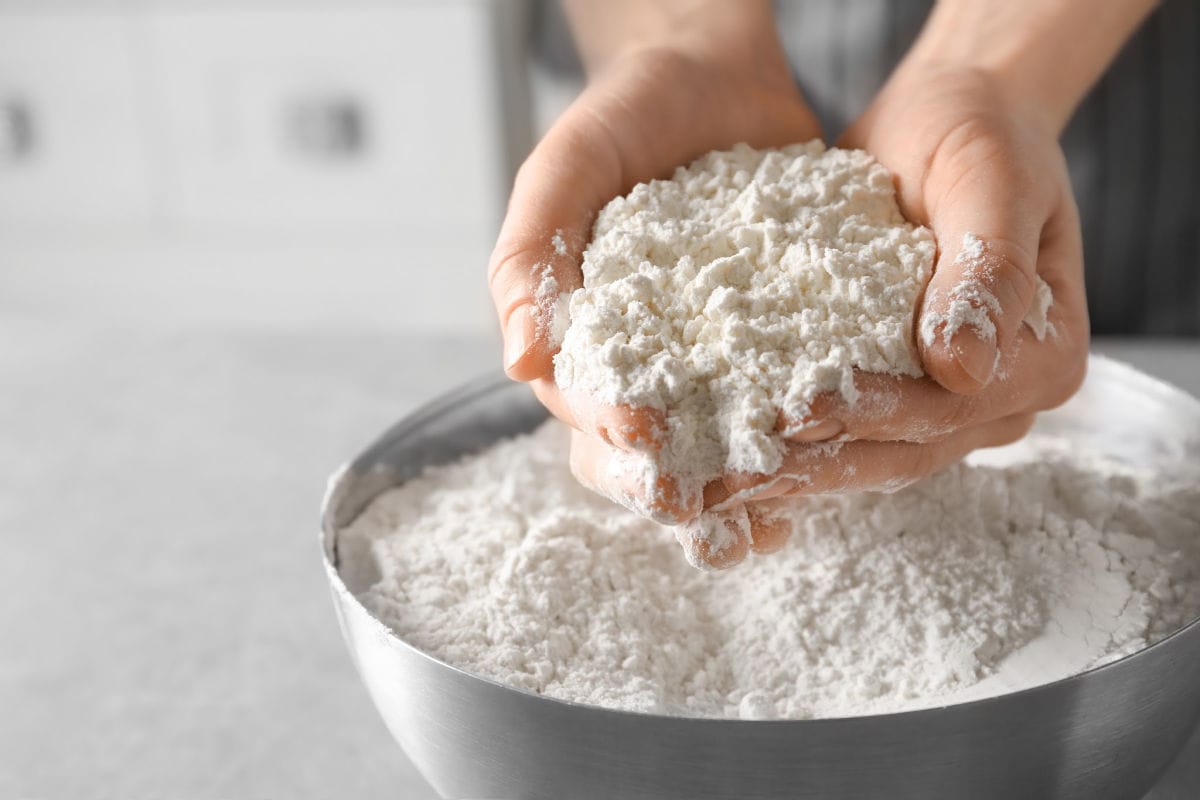 White flour in a silver bowl with hands scooping it up.