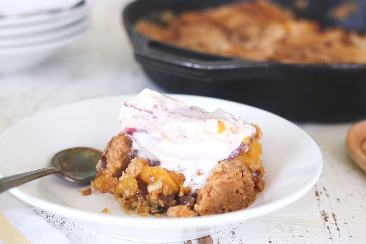 Serving of peach dump cake in a white bowl with ice cream melting on top.