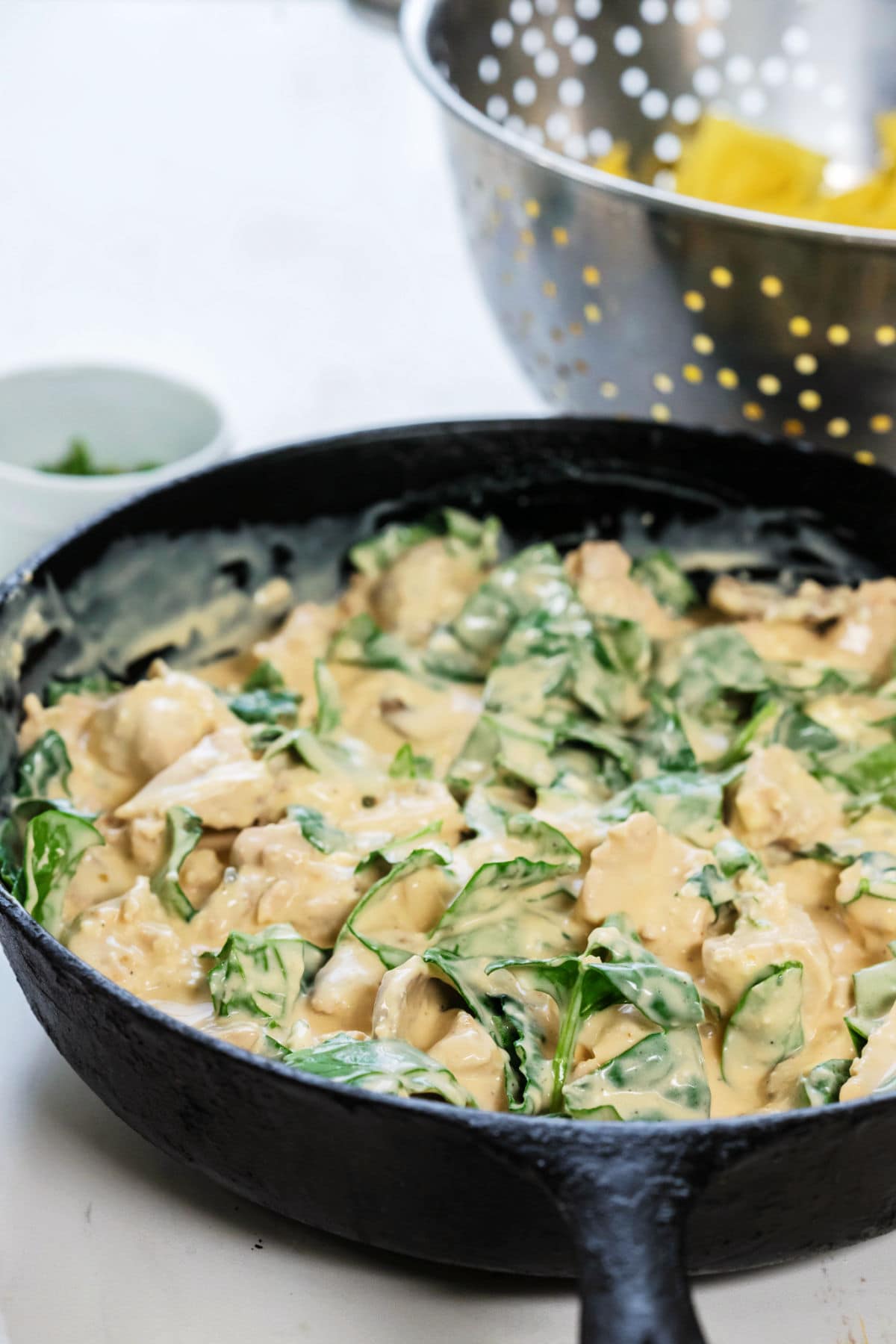 An iron skillet is full of the Alfredo chicken and spinach with cooked ravioli in a colander in the background.