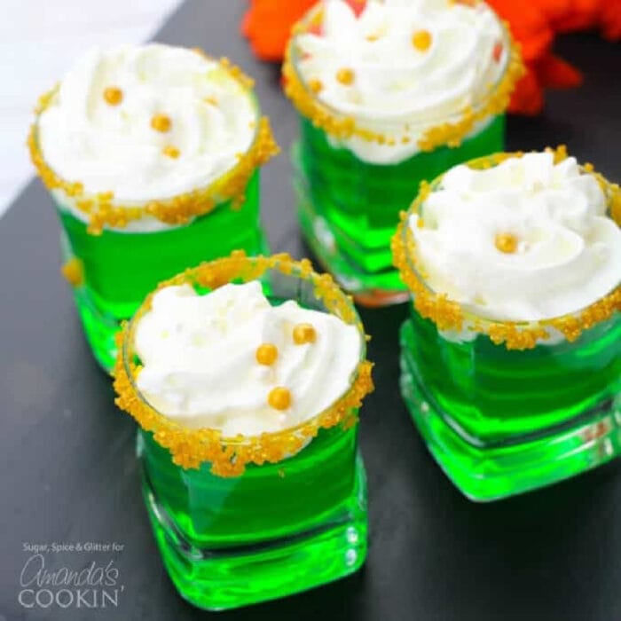 Four green jello shots with whipped cream on top in glasses garnished with gold sugar.