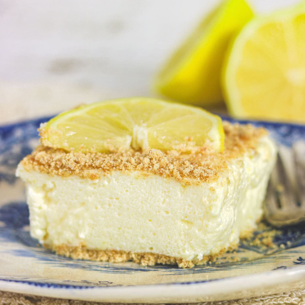 A square of lemon cheesecake on a flowered plate with a lemon slice on top.
