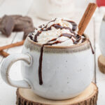 A cup of hot chocolate topped with whipped cream and garnished with chocolate sauce.