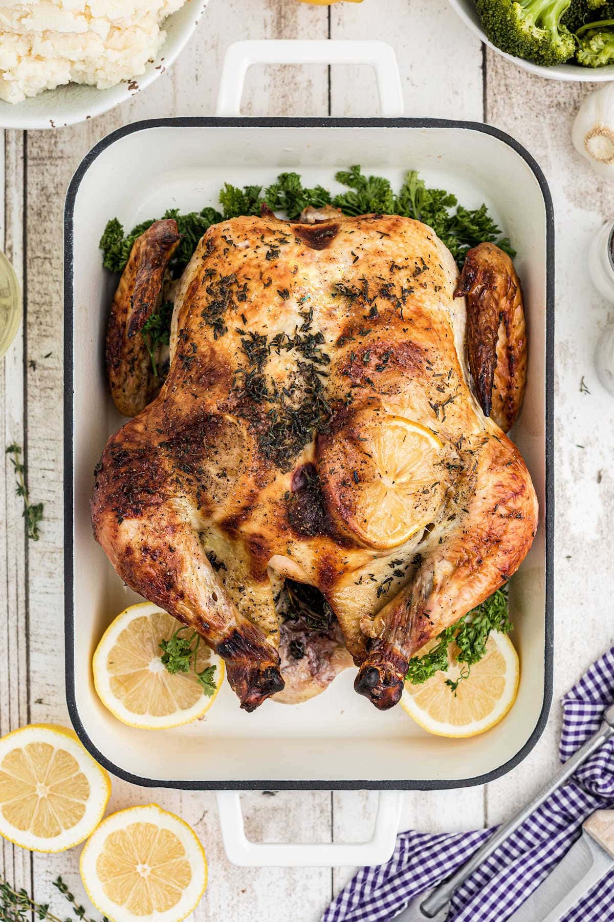 A roasting pan displays a cooked whole brined roast chicken adorned with slices of yellow lemon.