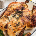 A whole roast chicken is displayed in a roasting pan; placed on a country white wooden table along with potatoes and broccoli sides and text.