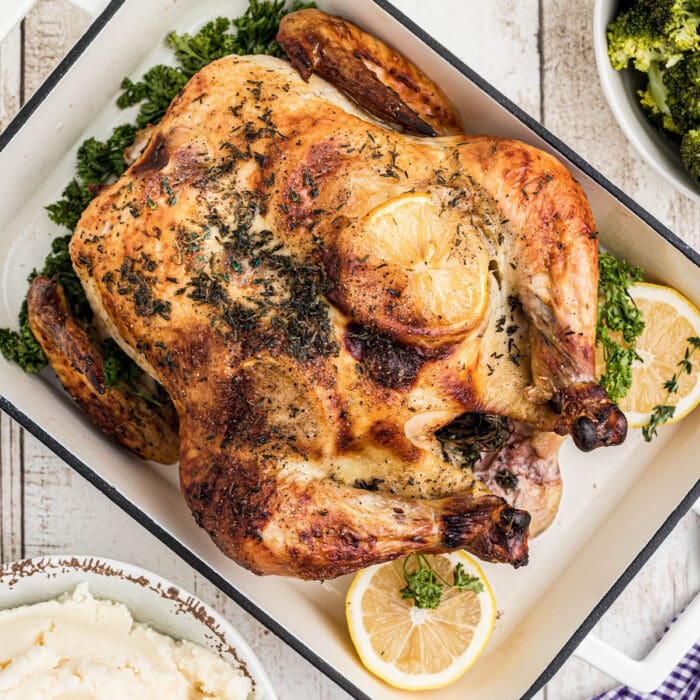 A whole brined roast chicken in a roasting pan with sliced lemons.