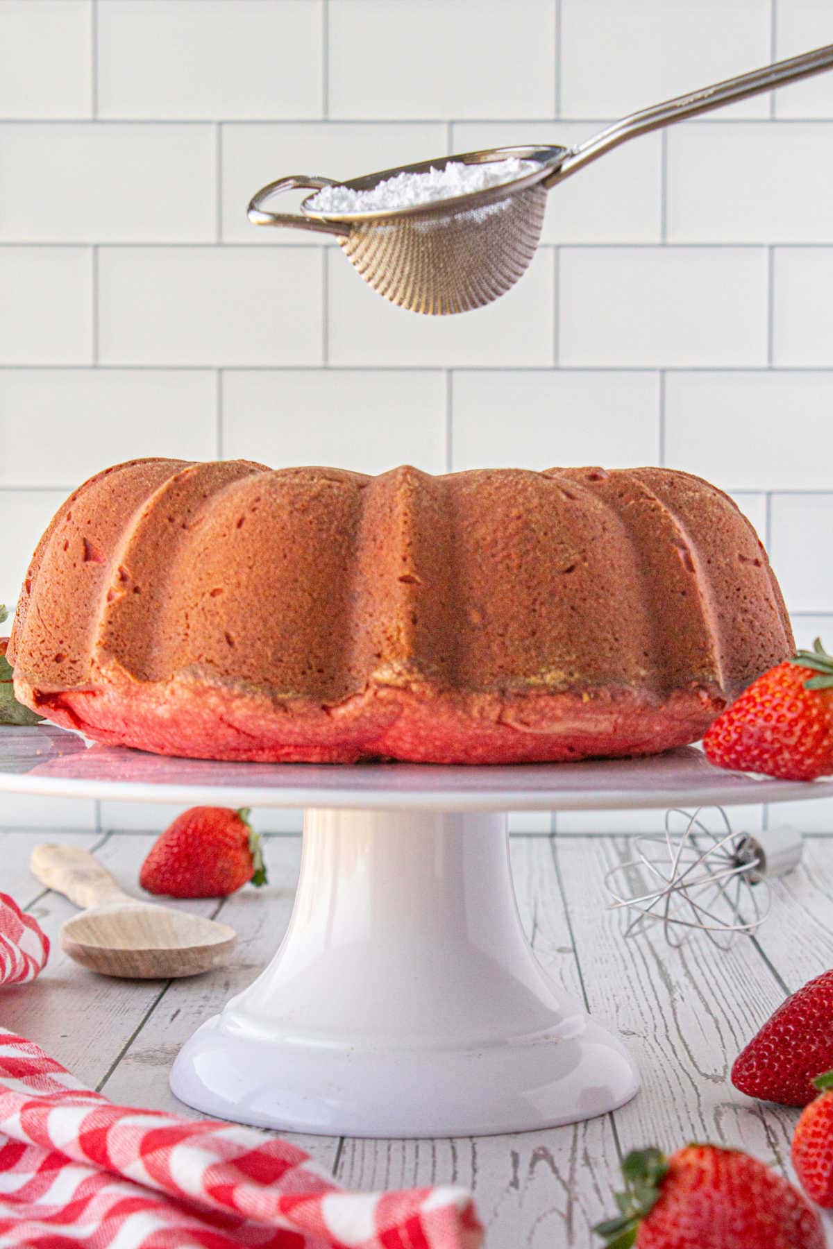 Baked strawberry bundt cake on a white cake stand as powdered sugar sifter is above cake ready to sprinkle on top.