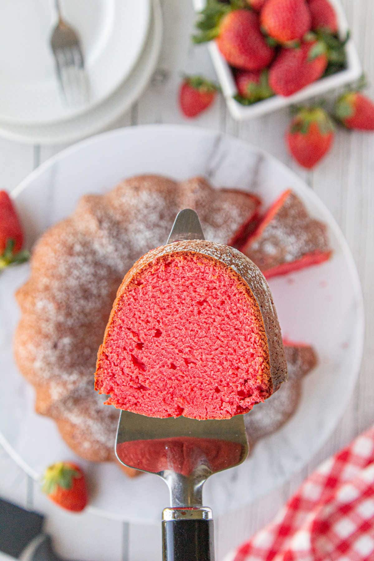 A cake server holds a slice of strawberry bundt cake over the sliced bundt cake and strawberries on marble counter and plate below.