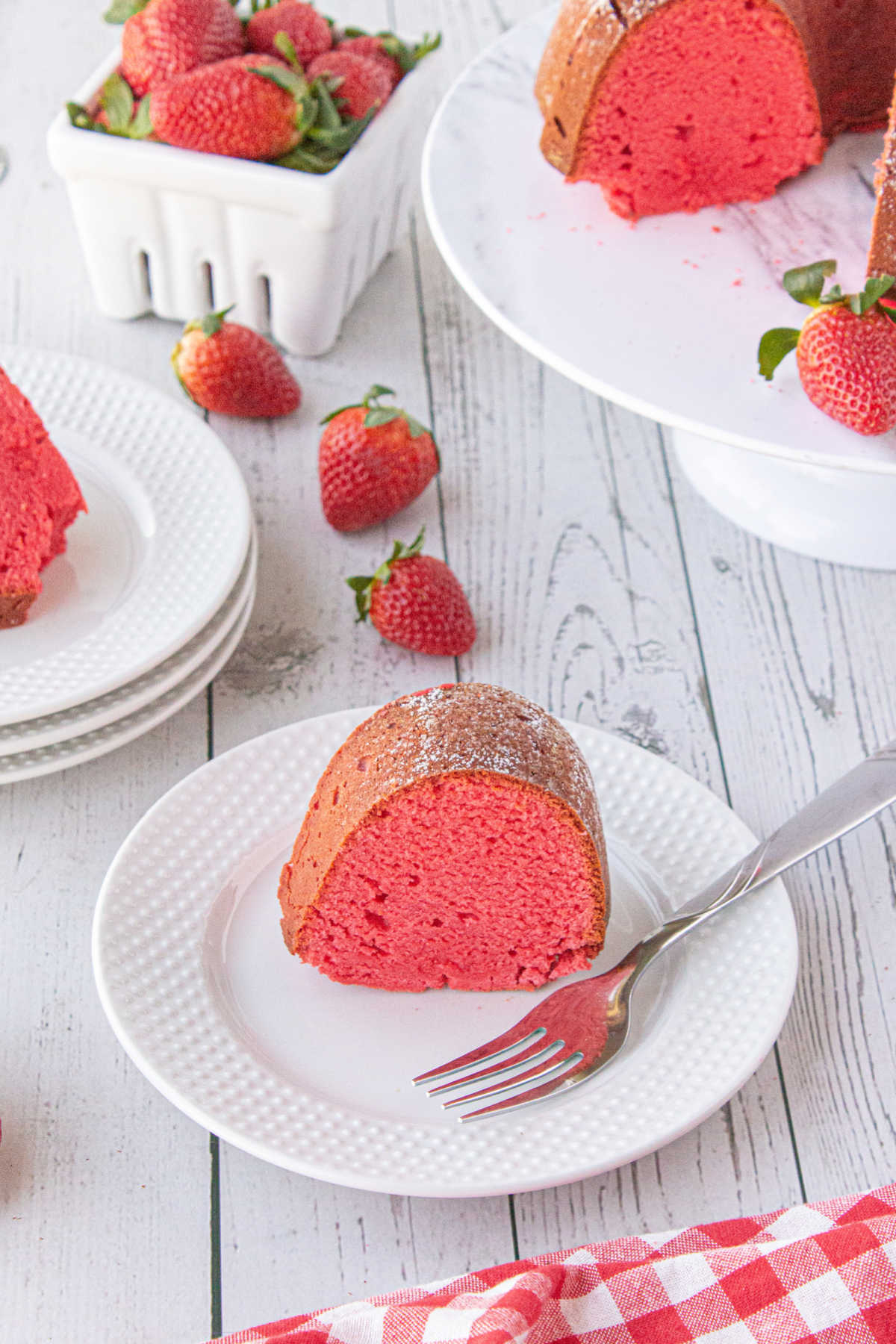 A piece of sliced red strawberry bundt cake on a white plate with a fork ready to serve as strawberries and cut cake are displayed behind it.