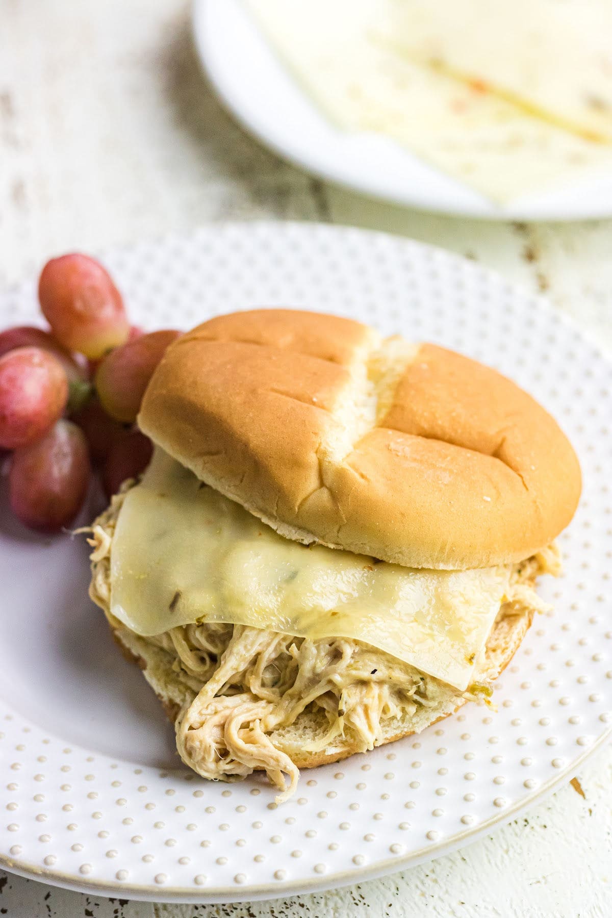 A shredded creamy ranch chicken sandwich with a slice of white cheese on a sandwich bun with a side of grapes.