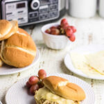 Slow Cooker Creamy Ranch Chicken Sandwich on a clean white plate with a side of purple grapes and Pin text.