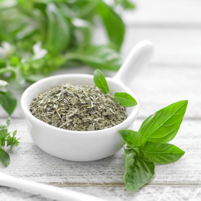 A white bowl of dried basil with fresh basil on the table surrounding it.