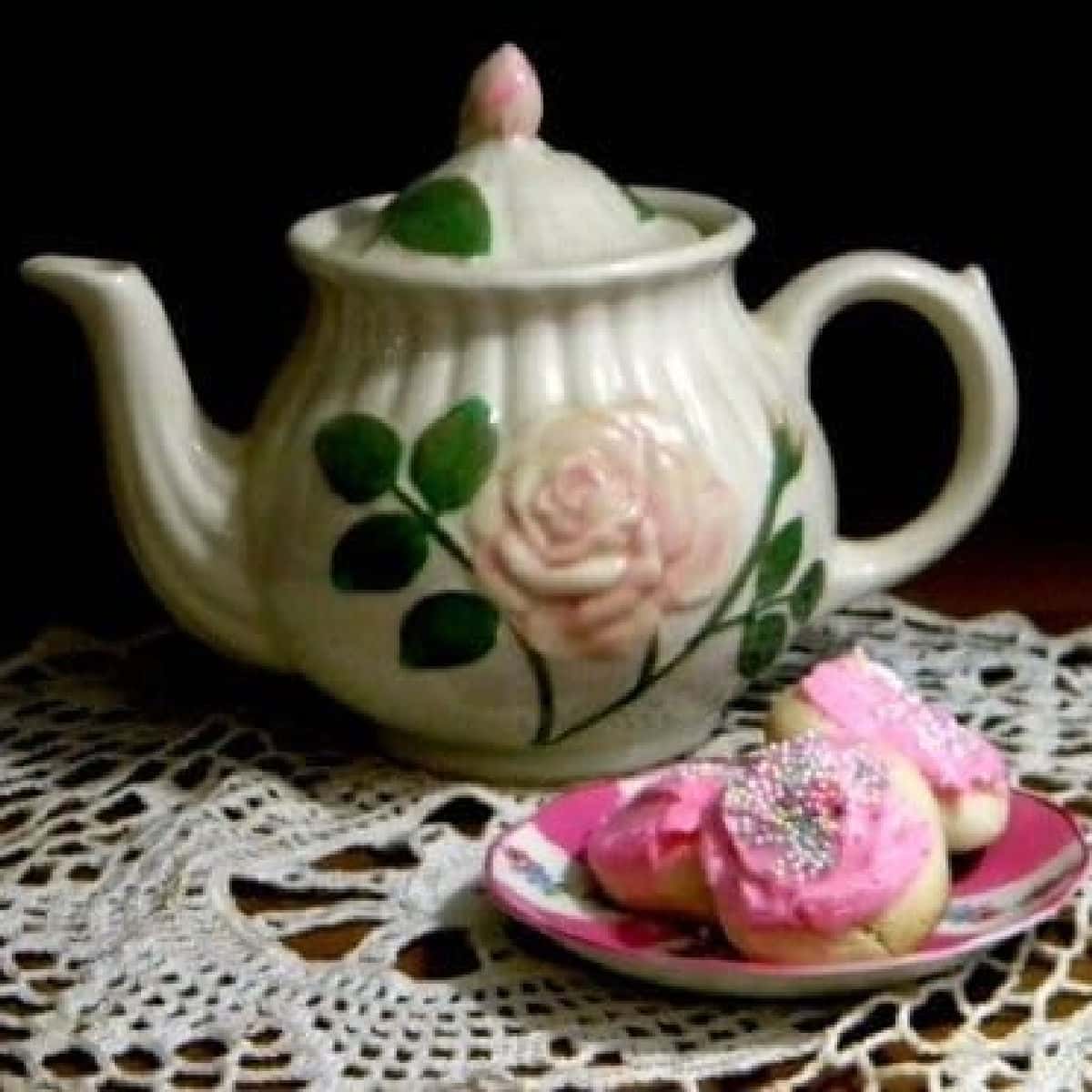 A white vintage teapot and a plate of cookies sit on a lace tablecloth.
