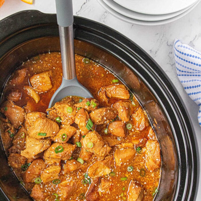 A crockpot full of orange chicken with a serving spoon in it.