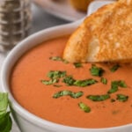 Closeup of a bowl of tomato soup with a grilled cheese sandwich being dipped in it.