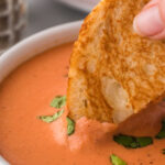 A toasted cheese sandwich being dipped into a bowl of tomato soup.