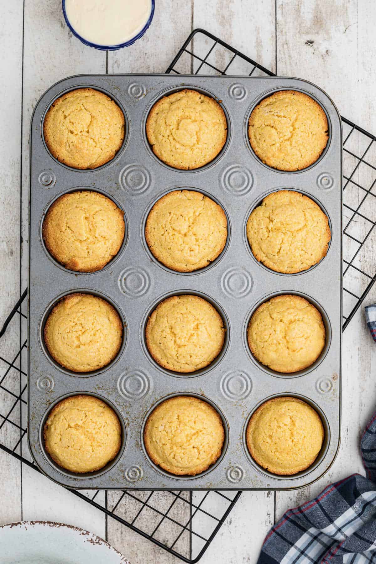 Overhead view of a muffin tin full of baked corn muffins cooling on a wire rack.